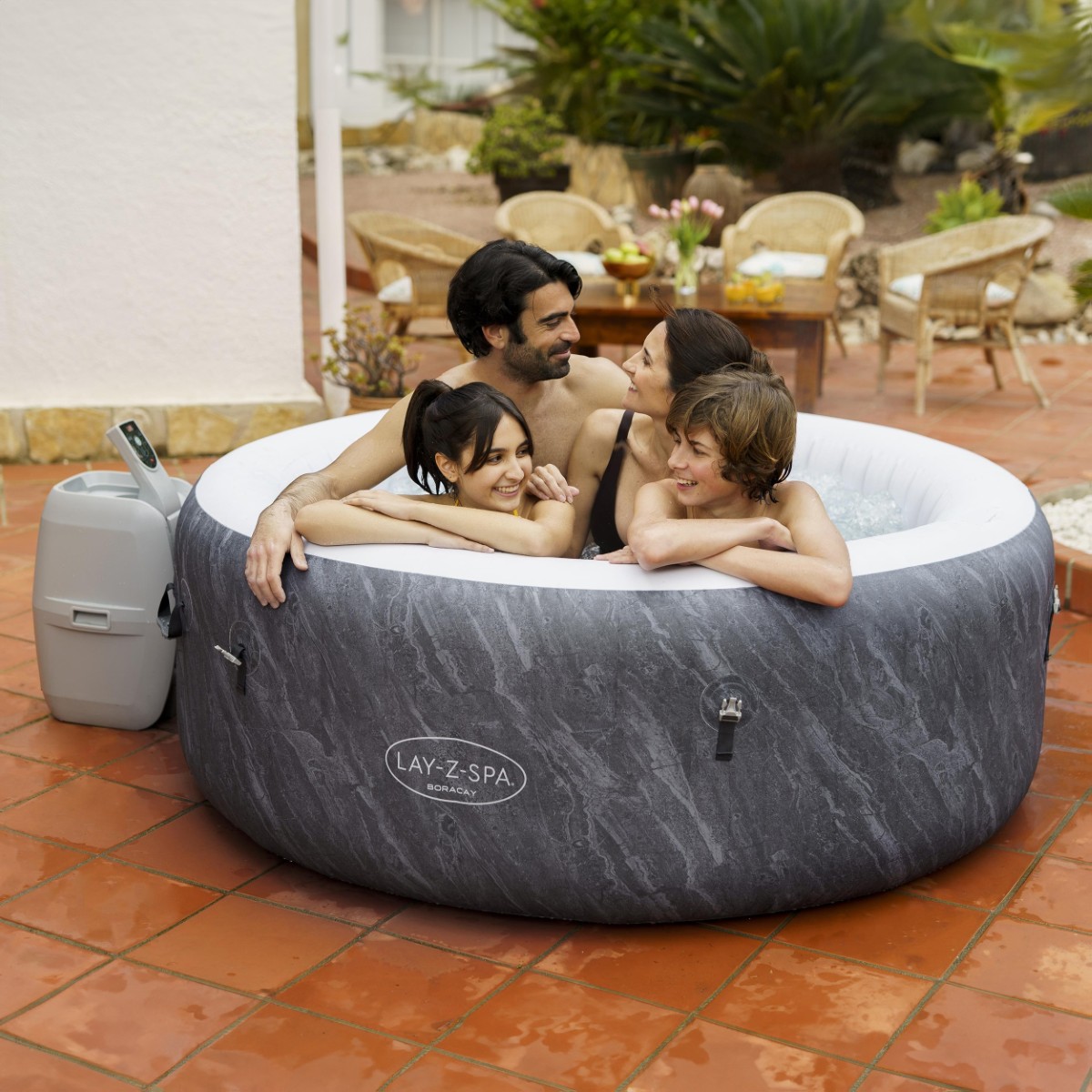 Czteroosobowa rodzina (rodzice i dwoje dzieci) relaksuje się w jacuzzi Lay-Z-Spa Boracay Smart ustawionym na tarasie. Jacuzzi ma ciemnoszare ścianki z efektem kamienia i białe wykończenie góry, idealnie komponując się z meblami ogrodowymi w tle.